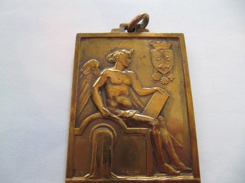 K&ouml;nnte beinhalten: Eine goldfarbene Metallmedaille mit einer gefl&uuml;gelten Figur, die eine Schriftrolle h&auml;lt. Die Medaille zeigt ein Wappen mit den Buchstaben "L.C." und einer Krone.