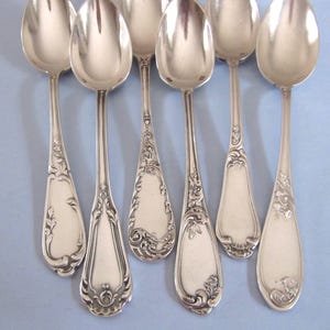 Puede incluir: Un conjunto de cucharas de plata con diseños florales y de volutas ornamentadas en los mangos y cuencos. Las cucharas están dispuestas sobre una superficie azul claro, mostrando sus superficies pulidas y reflectantes. Las cucharas son probablemente para servir o comer.