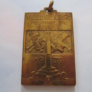 K&ouml;nnte beinhalten: Eine goldfarbene Metallmedaille mit dem Text "VILLE DE LIEGE FESTIVAL DE MUSIQUE 1939" und einem Design von Musikinstrumenten.