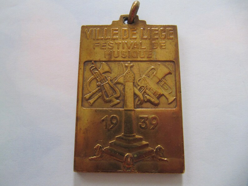 K&ouml;nnte beinhalten: Eine goldfarbene Metallmedaille mit dem Text "VILLE DE LIEGE FESTIVAL DE MUSIQUE 1939" und einem Design von Musikinstrumenten.