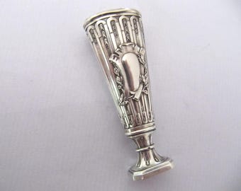 May include: A silver-toned antique wax seal with a decorative design. The seal features a shield-shaped emblem surrounded by laurel leaves and vertical lines. The base is rectangular.