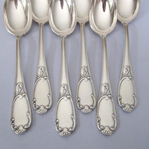 Puede incluir: Seis cucharas de plata con mangos ornamentados. Las cucharas tienen un acabado pulido y están dispuestas en fila.