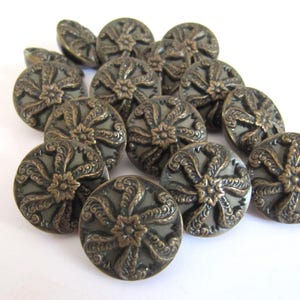 Op de afbeelding: Een verzameling ronde knopen met een donkergrijze metallic basis en een in reliëf aangebracht bronzen bloemmotief. Elke knoop heeft een centrale stervormige bloem omringd door zes gebogen, getextureerde bloemblaadjes in een cirkelvormig ontwerp.