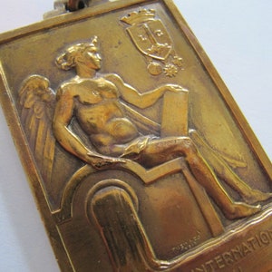 K&ouml;nnte beinhalten: Eine goldfarbene Metallmedaille mit einer gefl&uuml;gelten Figur, die eine Schriftrolle h&auml;lt. Die Medaille zeigt ein Wappen mit einer Krone und den Text "Internationale" unten.
