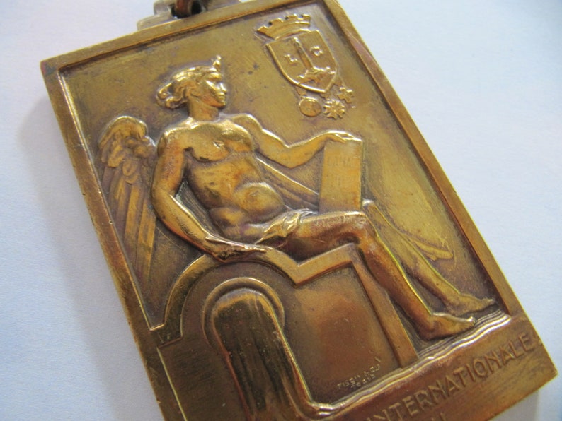 K&ouml;nnte beinhalten: Eine goldfarbene Metallmedaille mit einer gefl&uuml;gelten Figur, die eine Schriftrolle h&auml;lt. Die Medaille zeigt ein Wappen mit einer Krone und den Text "Internationale" unten.