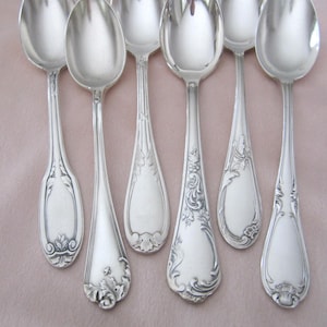 May include: A collection of silver spoons with ornate floral and scrollwork designs on the handles. The spoons are arranged on a light pink surface, showcasing their polished, reflective surfaces. The spoons vary in handle design.