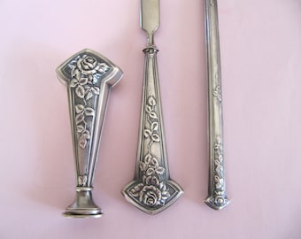 Op de afbeelding: Een set van drie zilverkleurige gereedschappen met bloemmotieven. De gereedschappen omvatten een briefopener, een zegellakzegel en een klein mes. De gereedschappen zijn allemaal versierd met rozen en bladeren.
