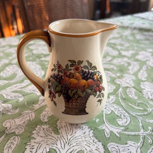 May include: Cream-colored ceramic pitcher with a brown handle and rim. The pitcher features a detailed illustration of a basket overflowing with various fruits and flowers. The pitcher is set on a green tablecloth with a white floral pattern.