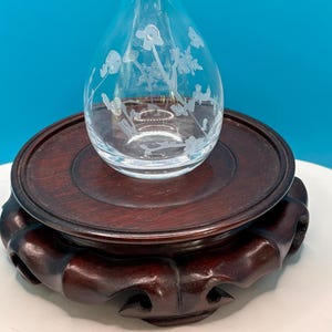 May include: A clear glass vase with etched floral designs sits on a dark brown wooden stand. The vase has a teardrop shape with a narrow neck. The stand is round with a carved base, showcasing a classic design.