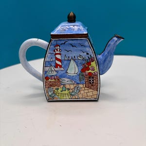 May include: A decorative ceramic teapot with a coastal scene. The teapot features a blue lid and spout, a white handle, and a square body. The body is painted with a lighthouse, sailboats, and a seaside cafe scene. The teapot is approximately 6 inches tall.