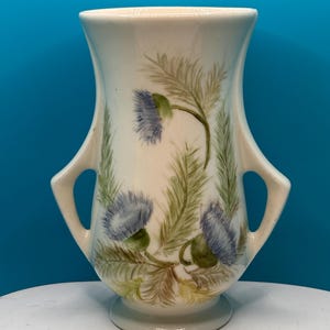 May include: A cream-colored ceramic vase with two side handles and a flared rim. The body of the vase is decorated with a hand-painted botanical pattern featuring blue thistle flowers and green foliage arranged around the center of the vessel.