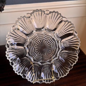 May include: A clear glass serving dish with a scalloped edge and a starburst design in the center. The dish is supported by a gold-colored metal stand. The dish is designed to hold multiple items.