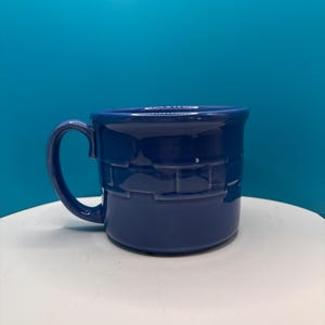 May include: A cobalt blue ceramic mug with a brick-like textured design around the base. The mug has a curved handle and a smooth, glossy finish. The mug is sitting on a white surface against a blue background.