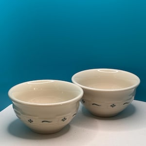 May include: Two off-white ceramic bowls featuring a subtle embossed grid pattern around the exterior. Each bowl is decorated with small, dark green floral accents and thin horizontal lines spaced evenly along the mid-section of the rounded body.