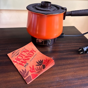 May include: A bright orange electric pot with a dark brown lid, handle, and base, featuring a West Bend brand label. Beside it lies a small orange booklet with the text West Bend 14 Cup Hot 'N Handy Pot Care/Use and Recipes printed in black.