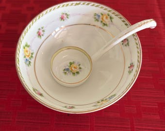 May include: A white porcelain soup bowl and ladle set with floral and gold trim. The bowl contains a smaller bowl with a matching floral design. The set is on a red surface.