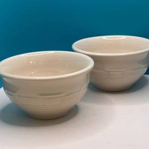 May include: Two off-white ceramic bowls with a smooth, glossy finish. Each bowl features a subtle, embossed basket-weave pattern around the exterior base. The bowls have a rounded shape with a slightly flared rim and a solid, uniform color.