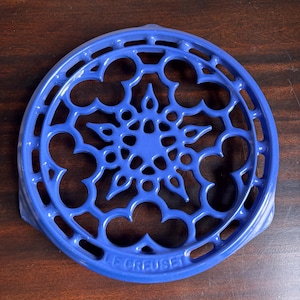 May include: A round, blue enameled cast iron trivet featuring an intricate, openwork geometric pattern. The design includes a central star-like motif surrounded by repeating clover-shaped cutouts. The brand name LE CREUSET is embossed.