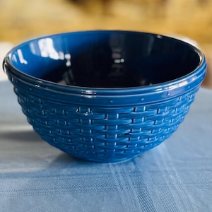 May include: A deep blue ceramic bowl with a woven basket design. The bowl has a smooth, glossy finish and is sitting on a light blue tablecloth. The bowl's interior is a darker shade of blue.