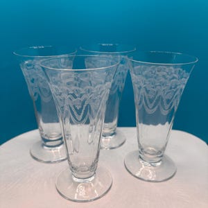 May include: Four clear, etched glass dessert dishes with a flared shape and a round base. The glasses have a delicate, repeating wave pattern around the upper portion. The background is a solid teal color.