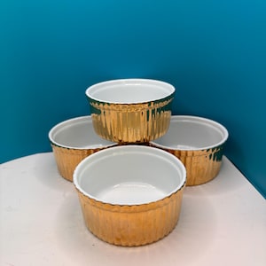 May include: Four gold-colored ramekins with a ribbed exterior and white interior. The ramekins are stacked in a pyramid shape, showcasing their design. The background is a solid teal color.