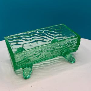 May include: A rectangular, light green glass dish with a textured, wood-grain-like pattern. The dish has four small feet and is filled with small bubbles. The glass is transparent, allowing light to pass through.