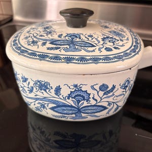 May include: A round, off-white enameled metal cooking pot with a matching lid. The exterior features a repeating blue floral and botanical pattern. A black, circular knob is centered on the lid. The pot has a white handle extending from the side.