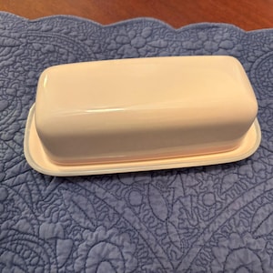 May include: A light pink butter dish with a matching lid. The rectangular dish sits on a white-rimmed base, and the lid is slightly rounded. The butter dish is on a blue quilted surface.