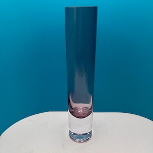 May include: A tall, slender cylindrical glass vase featuring a deep blue-tinted upper section that transitions into a clear, thick base with a rounded pink-tinted interior bubble detail at the bottom of the vessel's hollow center.