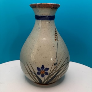 May include: A ceramic vase with a speckled, off-white body. It features a dark blue band near the neck and a brown rim. The vase is decorated with a blue flower and green grass design.