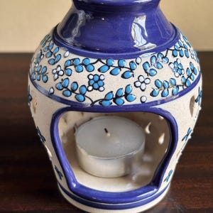 May include: A ceramic oil warmer with a glossy blue rim and base, featuring a cream-colored body decorated with hand-painted blue and white floral vines. The front has a large arched opening containing a small white tea light candle.