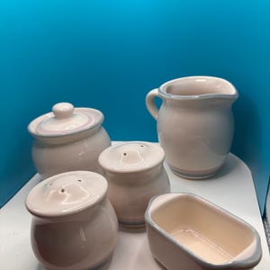 May include: A white ceramic kitchen set with a light blue and pink trim. The set includes a creamer, a covered sugar bowl, a butter dish, and two shakers. The pieces are arranged on a white surface against a blue background.