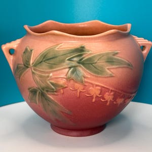 May include: A peach-colored ceramic vase with a ruffled rim and two handles. The vase features a raised design of green leaves and delicate floral accents. The vase is approximately 10 inches in diameter and 8 inches tall.
