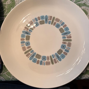 May include: White ceramic dinner plate with a decorative ring of geometric shapes in blue, brown, and gray. The plate has a wide rim and a clean, minimalist design, suitable for serving meals.
