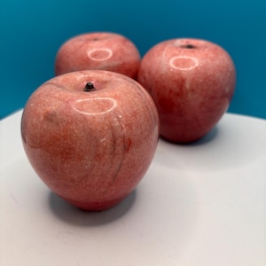 May include: Three decorative apples in shades of pink and red, with subtle marbling patterns. Each apple has a small, dark stem. The apples are arranged on a white surface against a blue backdrop.