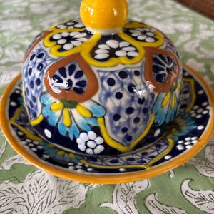 May include: A hand-painted ceramic butter dish with a lid and a matching plate. The dish features a vibrant design with blue, yellow, and white floral patterns on a dark blue background. The rim is a bright orange.