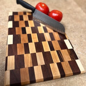 Könnte beinhalten: Ein Schachbrett-Schneidebrett aus hellem und dunklem Holz mit einem gezackten Messer und zwei roten Tomaten darauf.