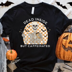 May include: Black t-shirt with a Halloween-themed graphic. The design features a skeleton holding a coffee cup, with the text "DEAD INSIDE BUT CAFFEINATED". The skeleton is set against a checkered orange and white background, with bats and stars.