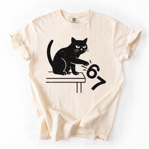 May include: Cream-colored t-shirt featuring a black cat design. The cat is sitting on a table and appears to be pushing the numbers "67" off the edge. The design has a distressed, vintage look.
