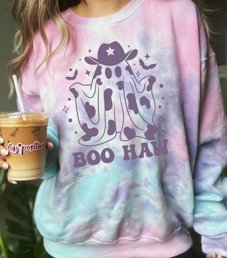 May include: A tie-dye sweatshirt with a purple ghost wearing a cowboy hat and the text "BOO HAW" in purple. The ghost has a cow print pattern.
