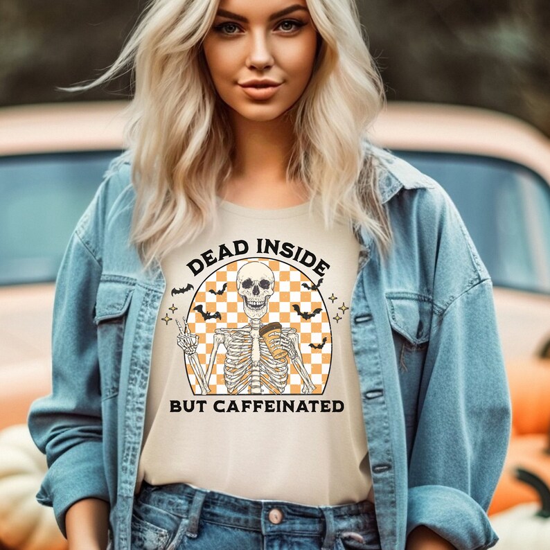 May include: Beige t-shirt with the text "DEAD INSIDE BUT CAFFEINATED" in a circular design. The design features a skeleton holding a coffee cup and giving a peace sign, with bats and stars. The shirt is worn by a person wearing a denim jacket.
