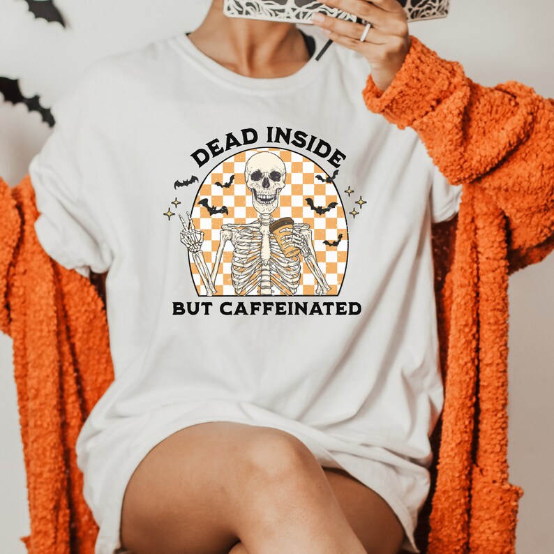 May include: White t-shirt with a Halloween-themed graphic. The design features a skeleton holding a coffee cup and giving a peace sign, with the text "DEAD INSIDE BUT CAFFEINATED" in a circular design with bats and stars.
