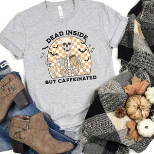 May include: A light gray t-shirt with the text "DEAD INSIDE BUT CAFFEINATED" above a skeleton graphic holding a coffee cup. The graphic includes bats and a checkered background. The shirt is paired with jeans and brown ankle boots.
