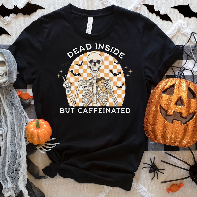 May include: Black t-shirt with a Halloween-themed graphic. The design features a skeleton holding a coffee cup, with the text "DEAD INSIDE BUT CAFFEINATED". The skeleton is set against a checkered orange and white background, with bats and stars.