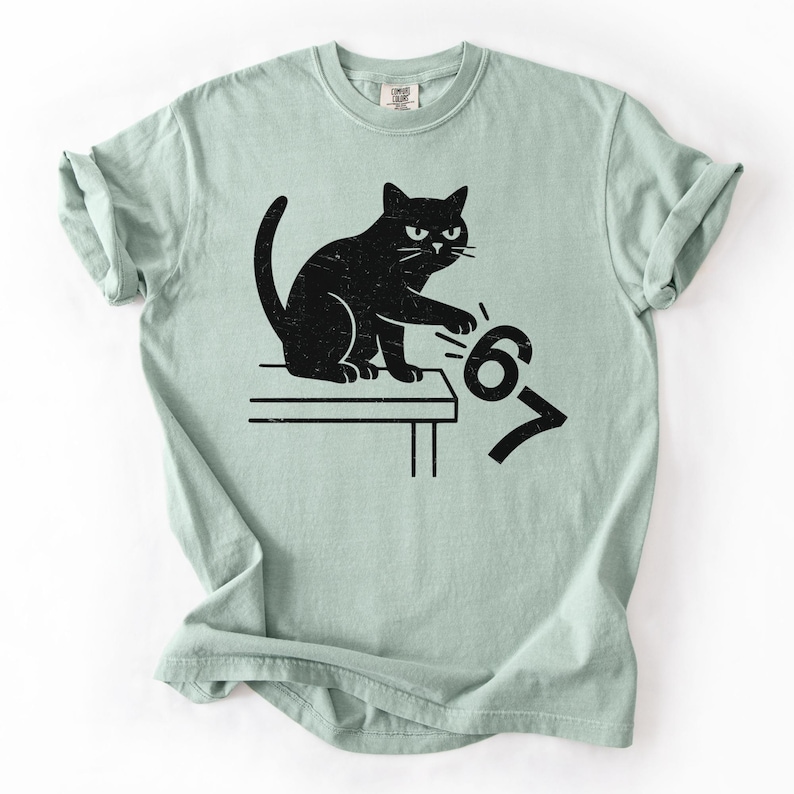 May include: A light green t-shirt featuring a black cat design. The cat is sitting on a table and appears to be pushing the numbers "6" and "7" off the table. The shirt has a crew neck and short sleeves.