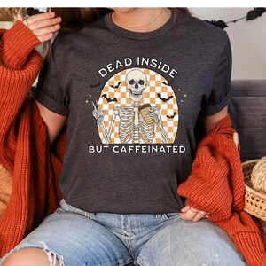 May include: Dark gray t-shirt with the text "DEAD INSIDE BUT CAFFEINATED" and a skeleton graphic. The skeleton is holding a coffee cup and making a peace sign. The design includes bats and an orange and white checkered background.