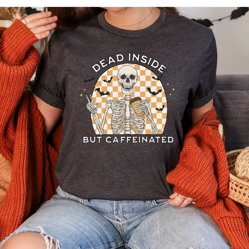 May include: Dark gray t-shirt with the text "DEAD INSIDE BUT CAFFEINATED" and a skeleton graphic. The skeleton is holding a coffee cup and making a peace sign. The design includes bats and an orange and white checkered background.