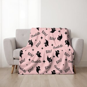 May include: A pink blanket with a repeating pattern of black ghosts, stars, and the word "BOO!" and the name "Charlotte". The blanket is draped over a light gray sofa.