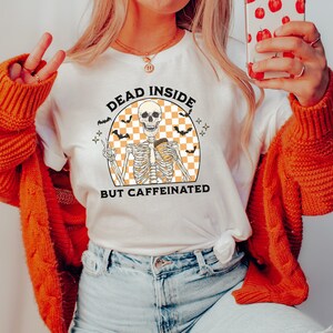 May include: White t-shirt with a skeleton graphic holding a coffee cup, surrounded by bats and the text "DEAD INSIDE BUT CAFFEINATED". The design features a checkered orange and white background. The shirt is part of a Halloween-themed collection.