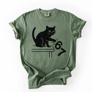 May include: Green t-shirt featuring a black cat illustration. The cat is sitting on a table and appears to be pushing the numbers "67" off the table. The shirt has a classic crew neck and short sleeves.